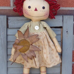 Op de afbeelding: Een ragdoll met rood garen haar, een groen-wit geblokte jurk en een bruine viltzonnebloem. De pop staat op een grijs houten oppervlak.
