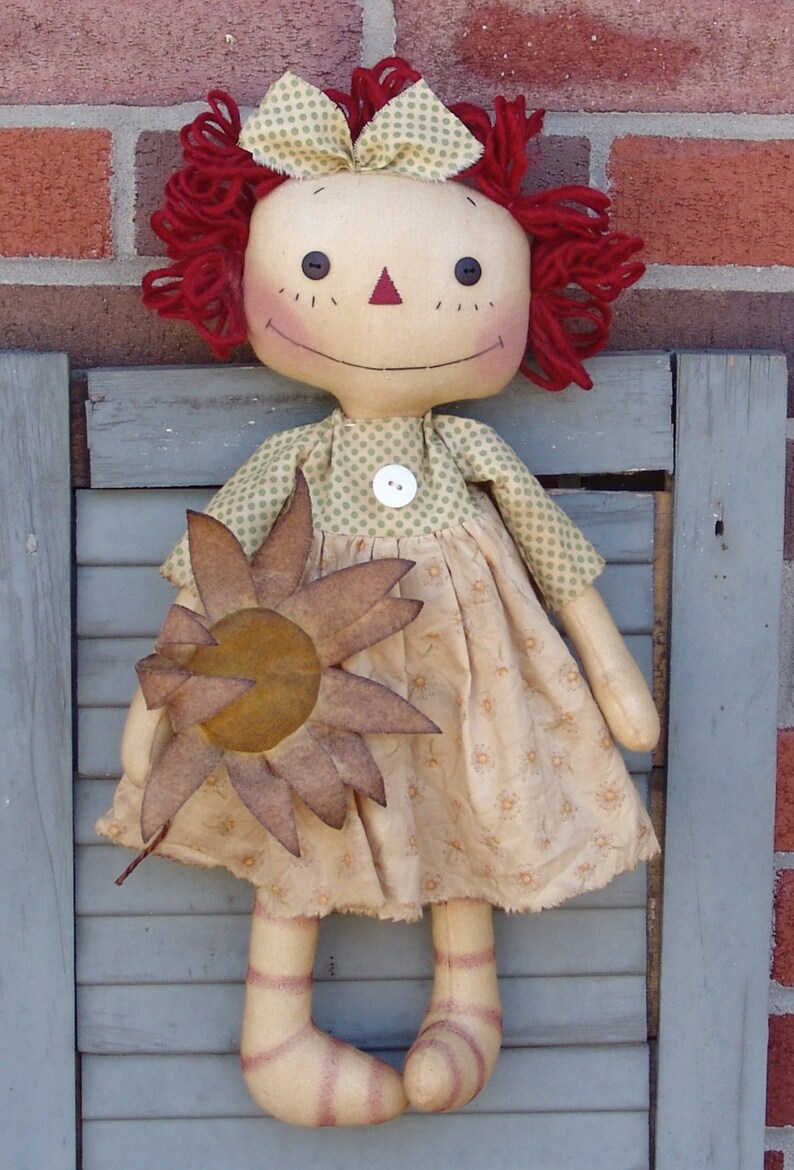 Op de afbeelding: Een ragdoll met rood garen haar, een groen-wit geblokte jurk en een bruine viltzonnebloem. De pop staat op een grijs houten oppervlak.
