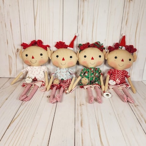 May include: Four handmade elf dolls with tan skin, red noses, and smiling faces. Each doll wears a different patterned dress and red and white striped legs. Two dolls have red bows, and two have Santa hats. The dolls are sitting on a light wood surface.