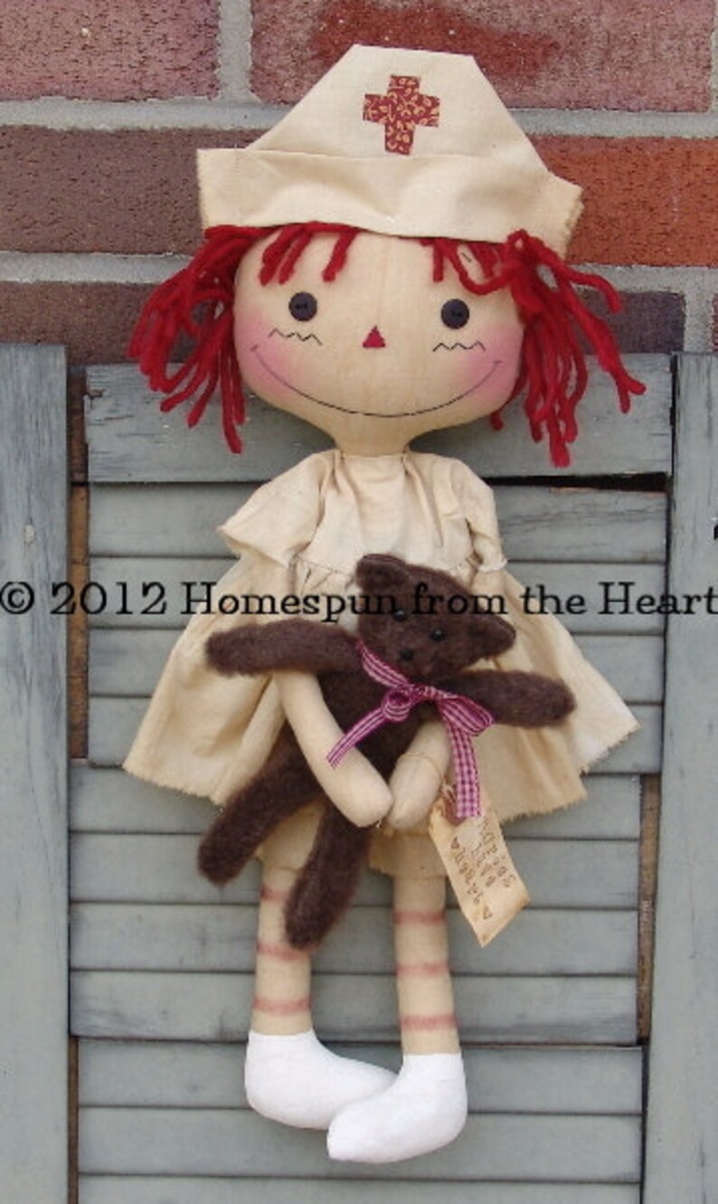 May include: A handmade doll dressed as a nurse, wearing a cream-colored uniform and cap with a red cross. The doll has red yarn hair and is holding a small brown teddy bear with a pink ribbon. The doll is approximately 18 inches tall.