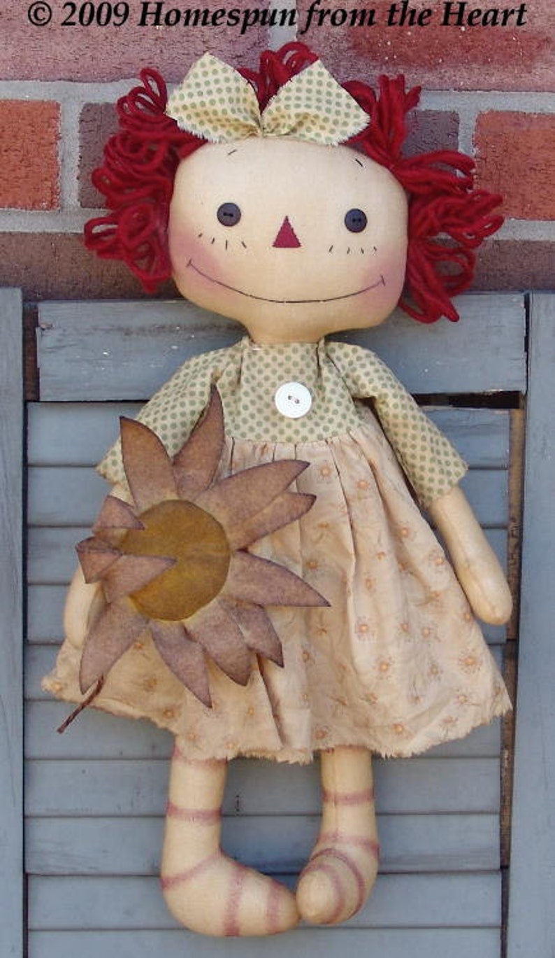 Op de afbeelding: Een handgemaakte lappop met rood garen haar, een groen-wit gebloemde jurk en een bruine viltbloem. De pop houdt de bloem in haar rechterhand. De pop zit op een houten oppervlak. &copy; 2009 Homespun from the Heart