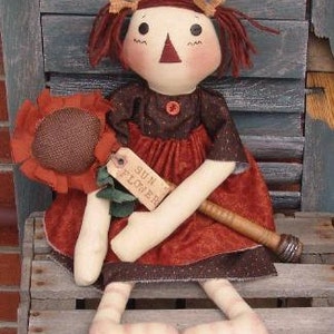 May include: A handmade rag doll with brown hair, wearing a brown and red dress, holding a brown stick with a sunflower attached. The doll is sitting on a wooden bench with a sign that says "Sun Flower".