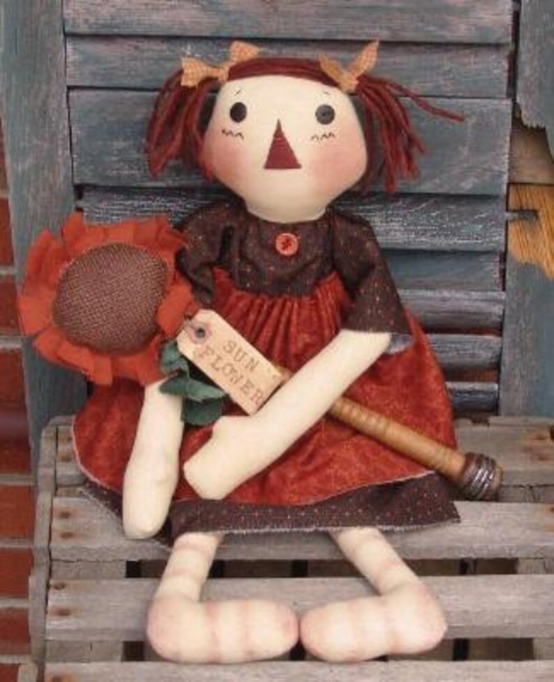 May include: A handmade rag doll with brown hair, wearing a brown and red dress, holding a brown stick with a sunflower attached. The doll is sitting on a wooden bench with a sign that says "Sun Flower".