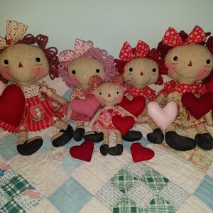 May include: Five handmade rag dolls with red and pink fabric hearts. The dolls are dressed in red and pink dresses with white polka dots. The dolls have red yarn hair and black button eyes.