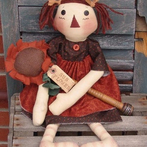 May include: A handmade rag doll with red hair, wearing a brown and red dress, holding a brown sunflower with a tag that says "SUN FLOWER".