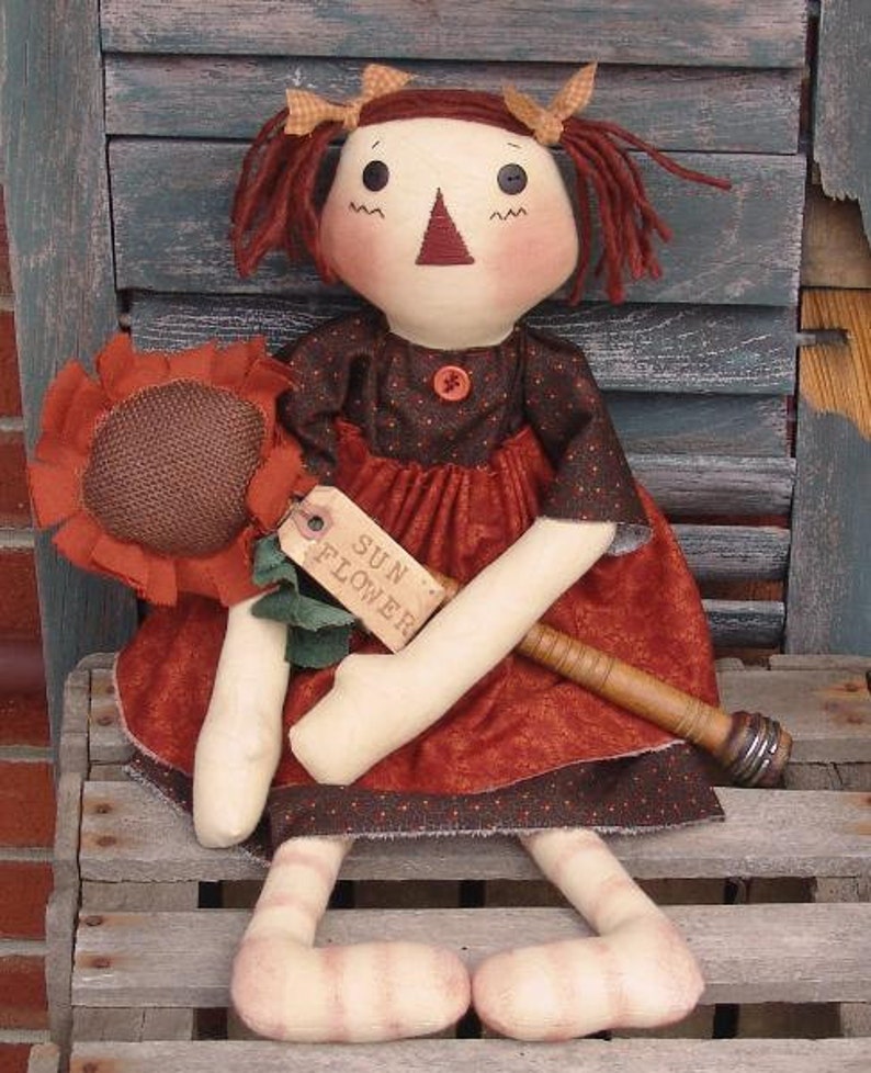 May include: A handmade rag doll with red hair, wearing a brown and red dress, holding a brown sunflower with a tag that says "SUN FLOWER".