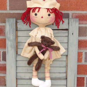 May include: A handmade fabric doll dressed as a nurse, wearing a cream-colored uniform and a white cap with a red cross. The doll has red yarn hair and is holding a small brown teddy bear with a pink ribbon. The doll is displayed against a weathered, blue-gray shutter.