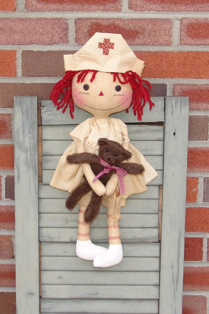 May include: A handmade fabric doll dressed as a nurse, wearing a cream-colored uniform and a white cap with a red cross. The doll has red yarn hair and is holding a small brown teddy bear with a pink ribbon. The doll is displayed against a weathered, blue-gray shutter.