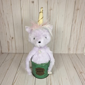 May include: A plush lavender unicorn with a cream-colored horn wrapped in gold thread, featuring a soft mane and a small unicorn charm necklace. The creature is seated inside a green metal tin labeled HELLICK MINTS with a small mint candy attached.