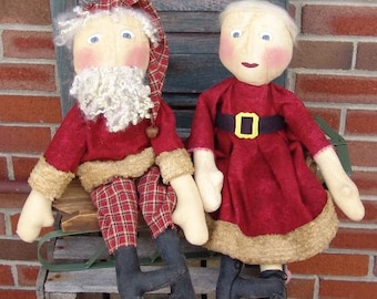 May include: Two handmade rag dolls, one dressed as Santa Claus and the other in a red dress with a gold belt. The Santa doll has a white beard and a red hat with white trim. The other doll has a red dress with a gold belt and a white fur trim.