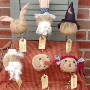 May include: Six handmade fabric doll heads on spring bases. The heads are of a bunny, a snowman, a witch, Santa Claus, a girl with red hair, and a man with a white beard and a blue and white hat. Each head has a tag with a name on it.