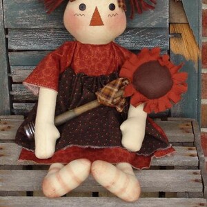 May include: A handmade rag doll with red yarn hair, a brown and white dress, and a brown sunflower in her hand.