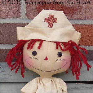 May include: A handmade rag doll with red yarn hair and a nurse's cap. The cap is cream-colored with a red cross design. The doll has button eyes, a stitched smile, and rosy cheeks. The doll is wearing a cream-colored outfit.