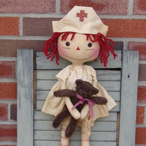 May include: A handmade doll dressed as a nurse, wearing a cream-colored cap with a red cross. The doll has red yarn hair and is holding a small brown teddy bear with a pink ribbon. The doll is standing in front of a brick wall.