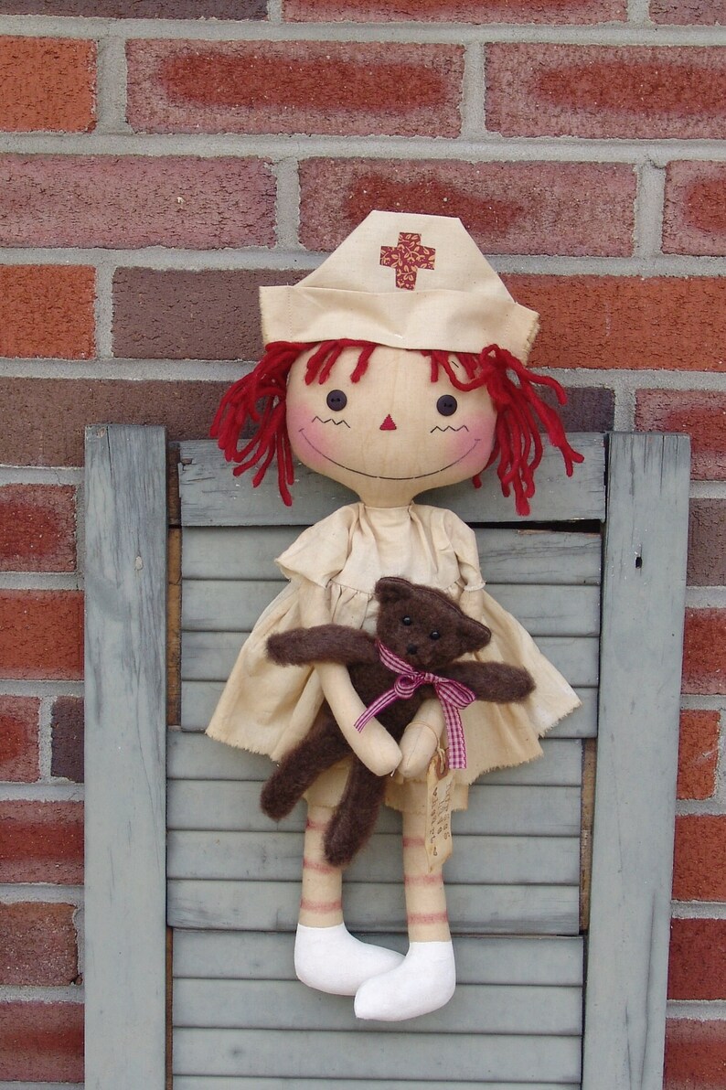 May include: A handmade doll dressed as a nurse, wearing a cream-colored cap with a red cross. The doll has red yarn hair and is holding a small brown teddy bear with a pink ribbon. The doll is standing in front of a brick wall.