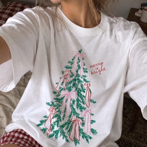 May include: A white short-sleeved t-shirt featuring a graphic of a green pine tree decorated with pink bows. The text "merry & bright" is printed in red cursive script to the right of the tree. The shirt has a crew neckline and a relaxed fit.