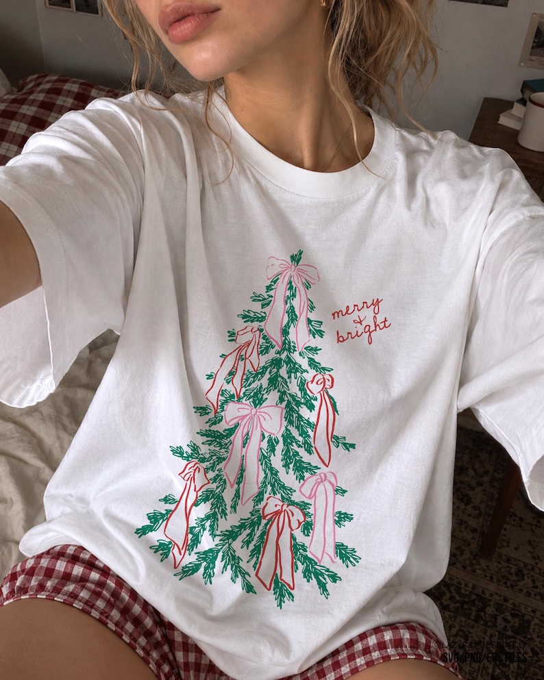 May include: A white short-sleeved t-shirt featuring a graphic of a green pine tree decorated with pink bows. The text "merry & bright" is printed in red cursive script to the right of the tree. The shirt has a crew neckline and a relaxed fit.
