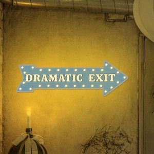 Puede incluir: Un letrero azul en forma de flecha con las palabras "DRAMATIC EXIT" en letras blancas. El letrero está iluminado con pequeñas luces y está montado en una pared. El letrero es parte de un diseño interior decorativo.