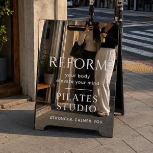 May include: A rectangular, mirrored A-frame sidewalk sign with a handle cutout at the top. The white text reads: REFORM your body elevate your mind PILATES STUDIO STRONGER. CALMER. YOU. The mirror reflects a street scene and two people standing.