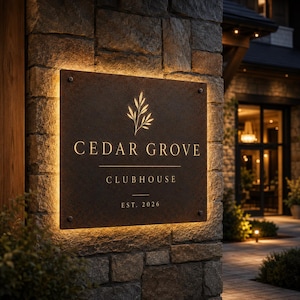 May include: A rectangular, dark, weathered metal sign mounted on a stone wall, featuring a backlit edge. The sign displays a gold botanical graphic above the text CEDAR GROVE, CLUBHOUSE, and EST. 2026 in a clean, serif font, all in gold.