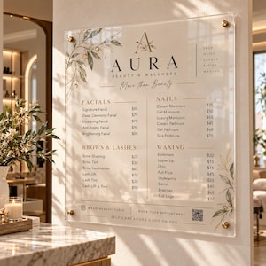 May include: A clear acrylic rectangular sign for Aura Beauty & Wellness featuring black text, a price list for facials, nails, brows, lashes, and waxing, and decorative botanical illustrations in the corners, mounted with gold hardware.