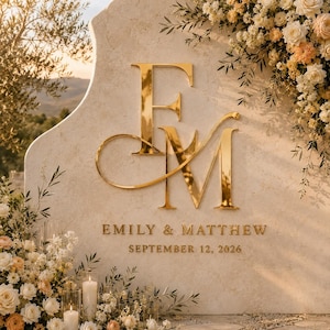 May include: A large, arched, beige stone sign featuring a reflective gold monogram of the letters F and M. Below the monogram, the text reads EMILY & MATTHEW and SEPTEMBER 12, 2026. The sign is decorated with white and peach flowers and greenery.