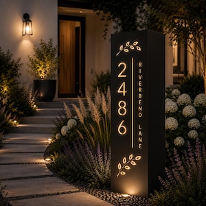 May include: A tall, rectangular, black metal house number sign with warm internal illumination. The sign displays the vertical numbers 2486, the text RIVERBEND LANE, and decorative leaf motifs at the top and bottom, all glowing from within.