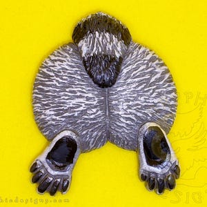 May include: A sculpted figurine depicting the rear view of a badger with textured gray and white fur, a black tail, and two front paws with black claws extended forward. The piece features a detailed, realistic fur pattern and glossy black accents.