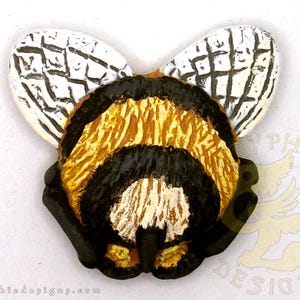 May include: A handcrafted bee-shaped decorative item. The bee features a detailed design with black, gold, and white colors. The wings are white with black vein-like patterns. The item is likely a brooch or a decorative element.