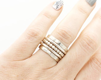 May include: A hand wearing a stack of five silver rings. The rings are different widths and have various textures, including hammered, beaded, and smooth.