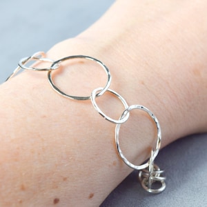 Puede incluir: Una pulsera de plata compuesta por múltiples eslabones circulares interconectados. Cada eslabón presenta una superficie martillada y texturizada que refleja la luz, creando una serie de anillos de distintos tamaños en cadena.