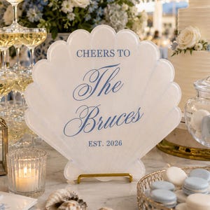 May include: A white, shell-shaped acrylic sign featuring blue text that reads CHEERS TO The Bruces EST. 2026. The sign is supported by a small gold metal stand and is positioned on a table among wedding decor including champagne glasses and macarons.