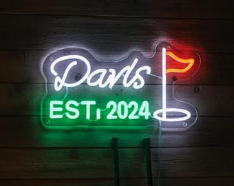 Puede incluir: Un letrero de neón con la palabra "Darts" en escritura blanca, "EST. 2024" en verde y una bandera de golf en rojo. Dos palos de golf están de pie. El letrero está montado en una pared de madera.