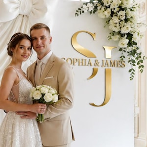 Op de afbeelding: Een bruiloftsachtergrond met een grote witte stoffen strik, witte bloemstukken en een gouden bord met de initialen SJ en de tekst SOPHIA & JAMES. Een persoon in een witte kanten jurk en een persoon in een beige pak staan voor het display.