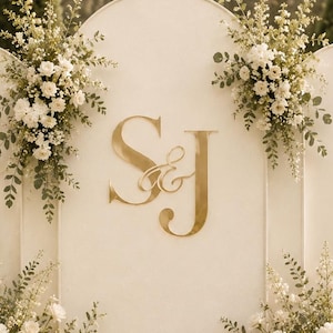 Può includere: Un grande fondale per matrimoni ad arco color bianco sporco con lettere S&J dorate al centro. La struttura è decorata con composizioni floreali bianche e verde, completata da lanterne dorate con candele accese alla base.