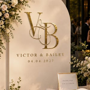 Può includere: Un grande cartello nuziale ad arco color crema con scritte dorate che recitano Victor & Bailey 04.04.2027 e un monogramma dorato VB. Il cartello è decorato con composizioni floreali bianche e si trova accanto a un tavolo con un libro degli ospiti.