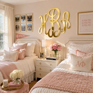Op de afbeelding: Een slaapkamer met twee eenpersoonsbedden met wit doorgestikt beddengoed en roze spreien. Elk bed heeft een sierkussen geborduurd met de Griekse letters Alpha Phi. Een groot gouden monogram in cursief schrift is aan de muur boven een nachtkastje.