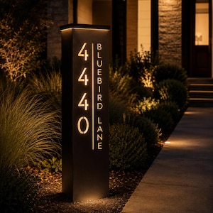 May include: A tall, rectangular, black metal address post featuring illuminated cut-out numbers 4440 and the text BLUEBIRD LANE vertically aligned. The structure emits a warm, golden glow from within, highlighting the typography against the dark finish.