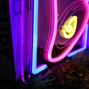May include: Neon sign with a pink, blue, and yellow design. The sign is shaped like a heart with a swirl inside. The sign is lit up and glowing.