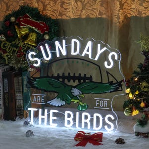 Może przedstawiać: Biały neonowy znak z napisem "Sundays Are For The Birds" i zielono-białą piłką nożną z zielonym orłem trzymającym piłkę nożną. Znak znajduje się na białym tle z czerwoną kokardką i zielonym drzewem bożonarodzeniowym w tle.