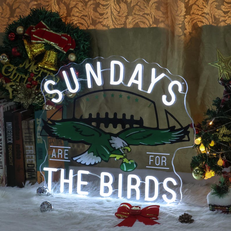 Może przedstawiać: Biały neonowy znak z napisem "Sundays Are For The Birds" i zielono-białą piłką nożną z zielonym orłem trzymającym piłkę nożną. Znak znajduje się na białym tle z czerwoną kokardką i zielonym drzewem bożonarodzeniowym w tle.