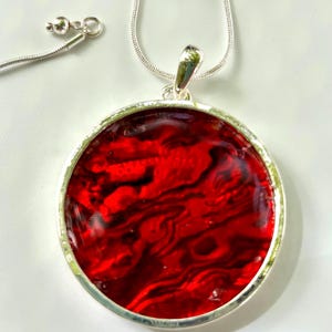 May include: A round pendant necklace featuring a vibrant red and black marbled pattern encased in a polished silver-toned metal frame. The pendant hangs from a matching silver-toned snake chain that includes a small spherical bead clasp detail.