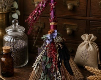 Puede incluir: Una escoba decorativa con un mango morado envuelto en hilo granate y adornado con flores secas, incluyendo ramas rojas y moradas, hojas de eucalipto verdes, plumas negras y un pequeño dije metálico circular con un diseño de estrella.
