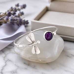 Könnte beinhalten: Ein silberfarbenes Armband mit einem Libellen-Design und einem runden, tiefvioletten Edelstein. Das offene Armband liegt auf einem weißen Stein, mit Lavendel und Band im Hintergrund. Schmuck.