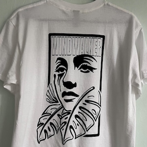Puede incluir: Camiseta blanca con un gráfico negro de un rostro estilizado, enmarcado por hojas, y la palabra "WINDWALKER" arriba. La camiseta está colgada en una percha negra contra una pared verde claro.