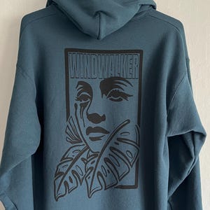 May include: Teal hoodie with a black graphic on the back. The graphic features a stylized face with a tear and tropical leaves, framed by the word "WINDWALKER". The hoodie has a hood and long sleeves.