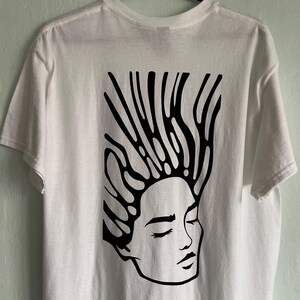 Puede incluir: Camiseta blanca con un gráfico negro de una cara con los ojos cerrados y cabello estilizado. La camiseta está colgada de una percha negra contra una pared verde claro. El diseño está centrado en la parte posterior de la camiseta.