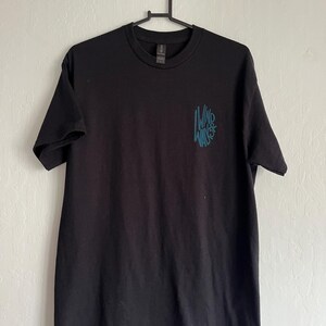 Puede incluir: Camiseta negra con el texto turquesa "Wind Walker" en un diseño estilizado en la parte delantera. La camiseta de manga corta está colgada en una percha negra contra una pared blanca. La camiseta está hecha de un material suave.