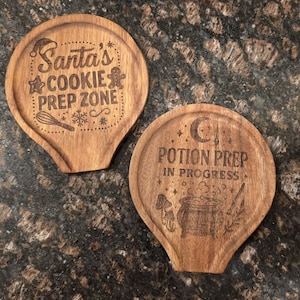 May include: Two wooden spoon rests with engraved text and designs. One says "Santa's COOKIE PREP ZONE" with a Santa hat, gingerbread man, and other festive icons. The other says "POTION PREP IN PROGRESS" with a cauldron and moon.
