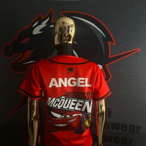 May include: Red baseball jersey with black trim, featuring the name "ANGEL" in white block letters. Below, a graphic of Lightning McQueen from the movie Cars is displayed. The jersey is on a gold mannequin against a dark background.